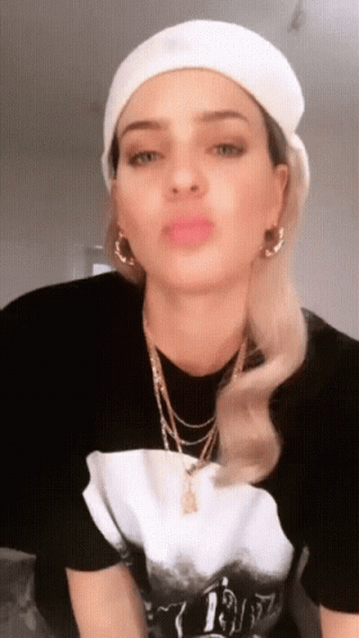 A Woman Wearing A White Hat And A Black T-Shirt With A Necklace That Says 1987 GIF