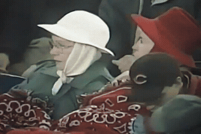 A Woman Wearing A White Hat And A Scarf Is Sitting In A Crowd Of People . GIF