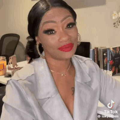 A Woman Wearing A White Jacket And Red Lipstick Is Sitting At A Table . GIF