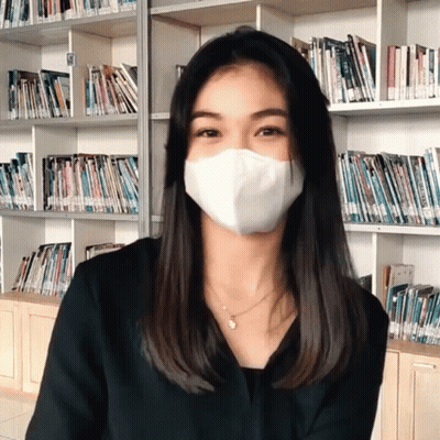 A Woman Wearing A White Mask Stands In Front Of A Book Shelf GIF