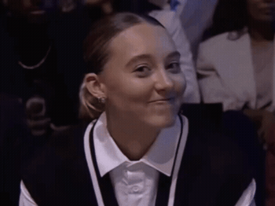 A Woman Wearing A White Shirt And A Black Vest Smiles While Sitting In A Crowd GIF