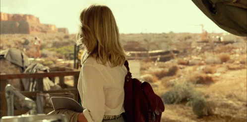A Woman Wearing A White Shirt And A Brown Bag Looks Out Over A Desert Landscape GIF