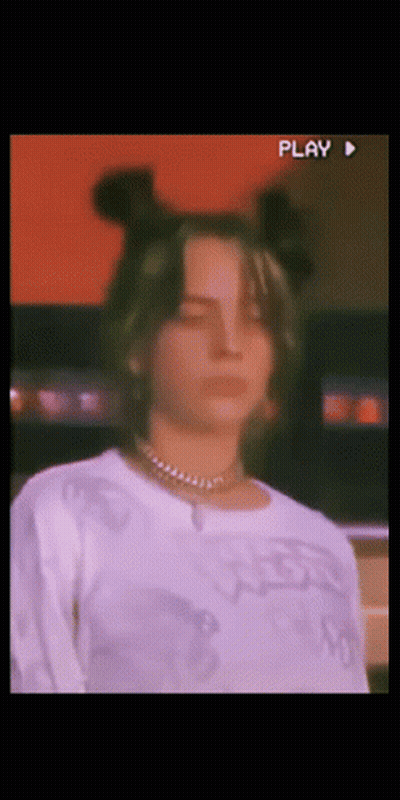 A Woman Wearing A White Shirt And A Necklace Looks At The Camera With A Play Button In The Corner GIF