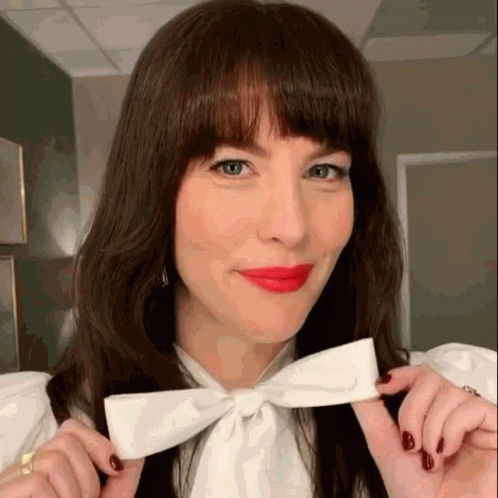 A Woman Wearing A White Shirt And Red Lipstick Is Holding A White Bow In Her Hand . GIF