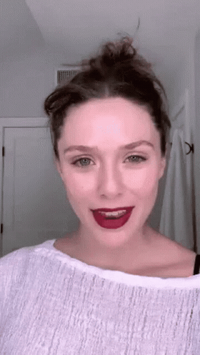 A Woman Wearing A White Sweater And Red Lipstick Is Standing In Front Of A Mirror . GIF