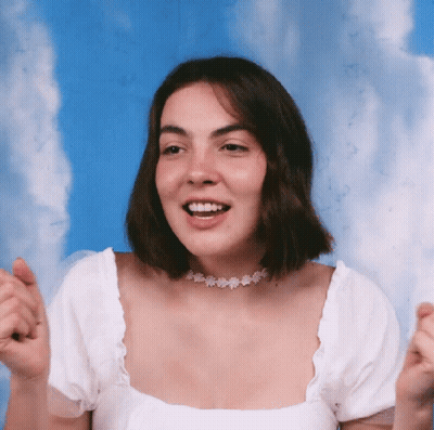 A Woman Wearing A White Top And A Choker Smiles With Her Hands In The Air GIF