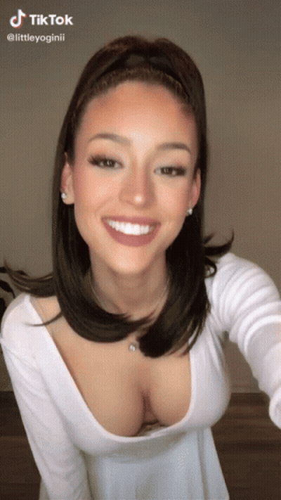 A Woman Wearing A White Top With A Plunging Neckline Is Smiling . GIF