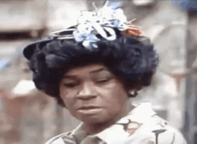 A Woman Wearing A Wig And A Hat With Flowers On It Is Making A Funny Face . GIF
