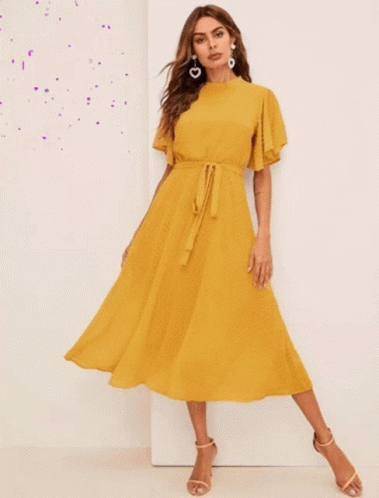 A Woman Wearing A Yellow Dress And Earrings Stands In Front Of A White Wall GIF