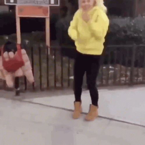 A Woman Wearing A Yellow Hoodie And Black Pants Is Standing On A Sidewalk . GIF