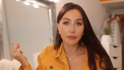 A Woman Wearing A Yellow Jacket And A Necklace Is Making A Funny Face . GIF