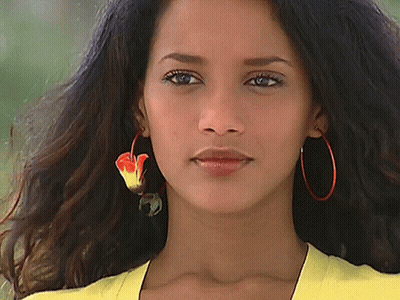 A Woman Wearing A Yellow Shirt And Red Hoop Earrings Looks At The Camera GIF