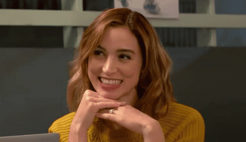 A Woman Wearing A Yellow Sweater Is Smiling With Her Hands On Her Chin GIF