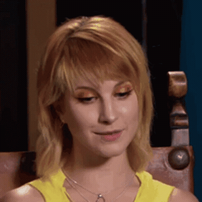 A Woman Wearing A Yellow Tank Top And A Necklace Is Sitting In A Chair And Smiling . GIF