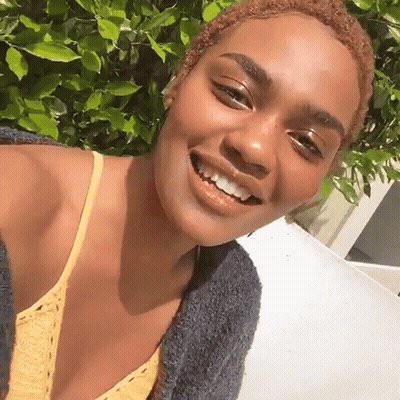 A Woman Wearing A Yellow Top And A Gray Sweater Smiles For The Camera GIF