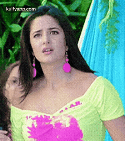 A Woman Wearing A Yellow Top And Pink Earrings Is Standing In Front Of A Blue Hammock . GIF