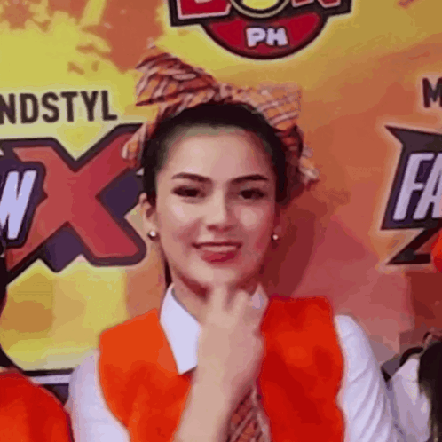 A Woman Wearing An Orange Vest And Tie Stands In Front Of A Ph Logo GIF
