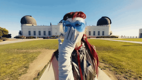 A Woman Wearing Blue Glasses And White Gloves Covering Her Face In Front Of A Building GIF