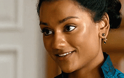 A Woman Wearing Earrings And A Blue Shirt Smiles GIF