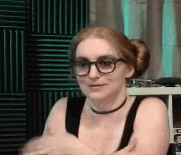 A Woman Wearing Glasses , A Choker , And A Bun Is Sitting In A Room . GIF