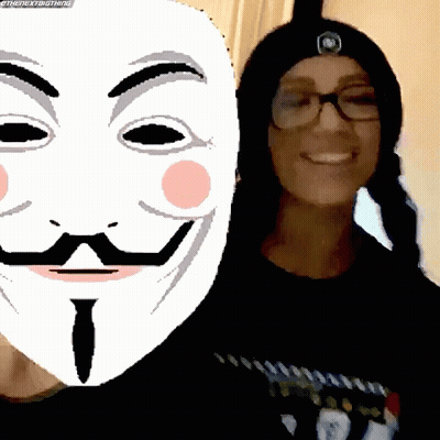 A Woman Wearing Glasses And A Beanie Stands Next To A Mask With A Mustache And Eyebrows GIF