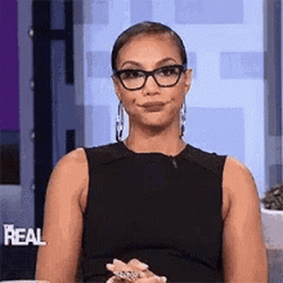 A Woman Wearing Glasses And A Black Dress Is Sitting On A Couch . GIF