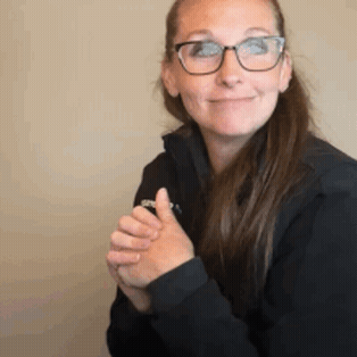 A Woman Wearing Glasses And A Black Jacket Is Smiling While Holding Her Hands Together . GIF
