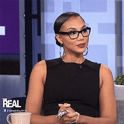 A Woman Wearing Glasses And A Black Tank Top Is Sitting In Front Of A Sign That Says Real GIF