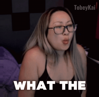 A Woman Wearing Glasses And A Black Top Says What The GIF