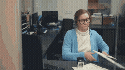 A Woman Wearing Glasses And A Blue Cardigan Is Sitting At A Desk GIF