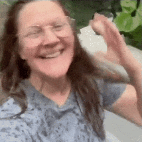 A Woman Wearing Glasses And A Blue Shirt Is Smiling And Waving Her Hands . GIF