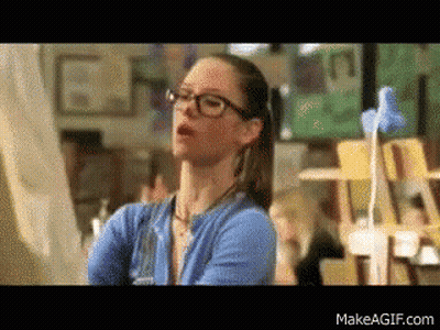 A Woman Wearing Glasses And A Blue Sweater Is Standing In Front Of A Painting GIF