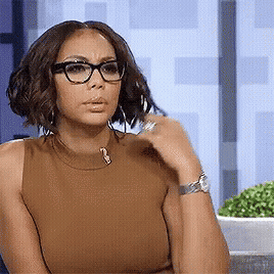 A Woman Wearing Glasses And A Brown Tank Top Is Sitting Down And Looking At The Camera . GIF