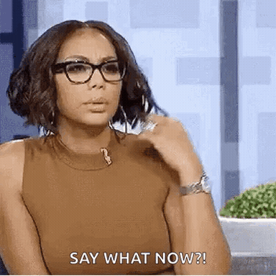A Woman Wearing Glasses And A Brown Tank Top Is Sitting Down And Saying `` Say What Now '' . GIF