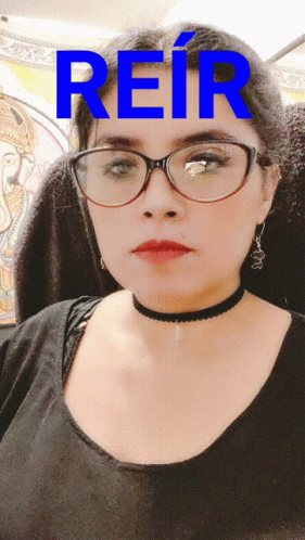 A Woman Wearing Glasses And A Choker Stands In Front Of A Blue Sign That Says Reir GIF