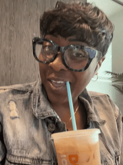 A Woman Wearing Glasses And A Denim Jacket Drinks Through A Straw From A Cup That Has The Letter D On It GIF