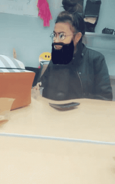 A Woman Wearing Glasses And A Fake Beard Sits At A Table GIF