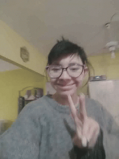 A Woman Wearing Glasses And A Gray Jacket Is Making A Peace Sign GIF