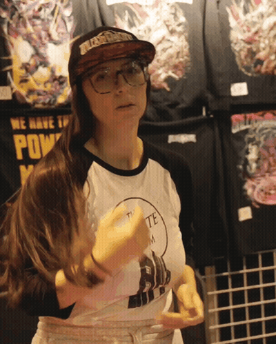 A Woman Wearing Glasses And A Hat Stands In Front Of A Sign That Says ' We Have The Power ' GIF