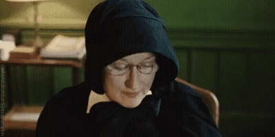 A Woman Wearing Glasses And A Hood Is Sitting At A Desk . GIF