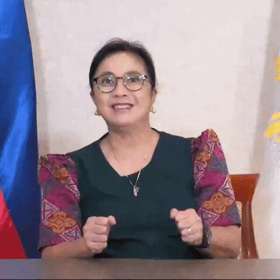 A Woman Wearing Glasses And A Necklace Is Sitting At A Desk With Her Hands In The Air . GIF