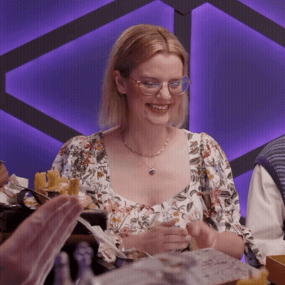 A Woman Wearing Glasses And A Necklace Smiles At A Table GIF