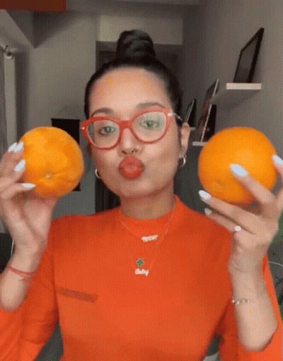 A Woman Wearing Glasses And A Necklace That Says Baby Holds Two Oranges GIF