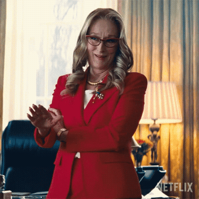 A Woman Wearing Glasses And A Red Suit Is Standing In Front Of A Netflix Sign GIF