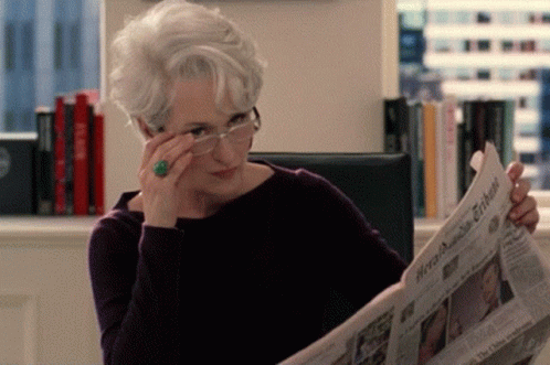 A Woman Wearing Glasses And A Ring Is Reading A Newspaper Called The New York Times GIF
