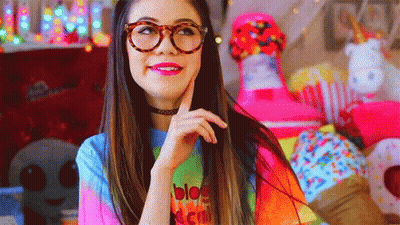 A Woman Wearing Glasses And A Tie Dye Shirt Is Standing In Front Of A Pile Of Stuffed Animals . GIF