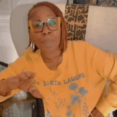 A Woman Wearing Glasses And A Yellow Sweater Is Sitting In A Chair And Making A Funny Face . GIF