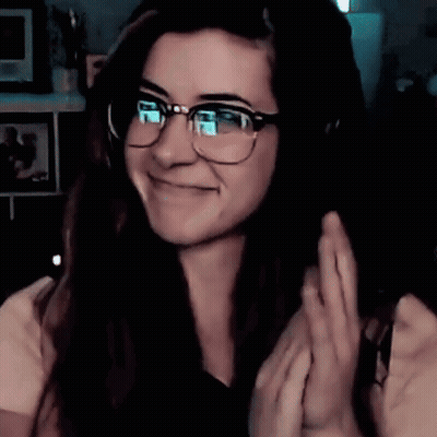 A Woman Wearing Glasses And Headphones Is Smiling And Waving GIF