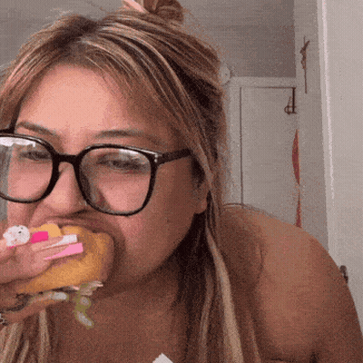 A Woman Wearing Glasses Is Eating A Hamburger With A Hello Kitty On It GIF