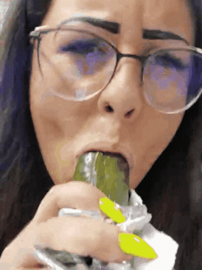 A Woman Wearing Glasses Is Eating A Pickle With Her Tongue GIF